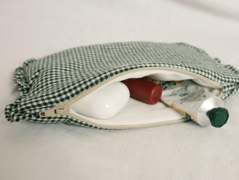 May include: A green and white gingham cosmetic bag with a beige zip, open to reveal a white bar of soap, a red tube, and a tube of cream. The bag is on a white surface.