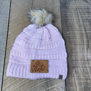 Puede incluir: Un gorro de punto rosa con un pompón de piel sintética en la parte superior. El gorro tiene un parche de cuero marrón con el texto "Let it snow" en blanco.