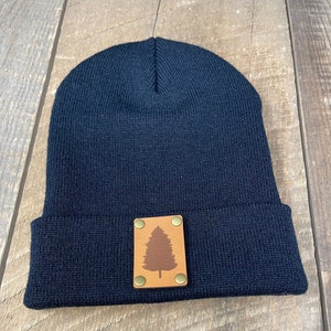 May include: Black knit beanie with a brown leather patch featuring a silhouette of a pine tree. The patch has four gold metal rivets.