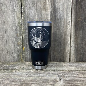 May include: Black Yeti tumbler with a laser engraved image of a white-tailed deer in a forest setting. The tumbler has the word "YETI" engraved on the bottom.