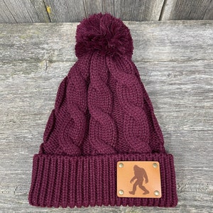 Könnte beinhalten: Eine bordeauxfarbene Strickmütze mit einem Bommel oben und einem braunen Lederpatch mit einer Bigfoot-Silhouette auf der Vorderseite.