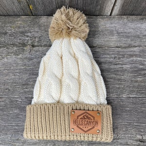 Könnte beinhalten: Eine weiße und braune Strickmütze mit einem Bommel oben. Die Mütze hat einen braunen Lederpatch mit dem Text "HELLS CANYON" und einer Berggrafik.