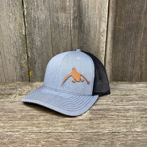 May include: A gray and black trucker hat with a brown leather duck silhouette on the front.