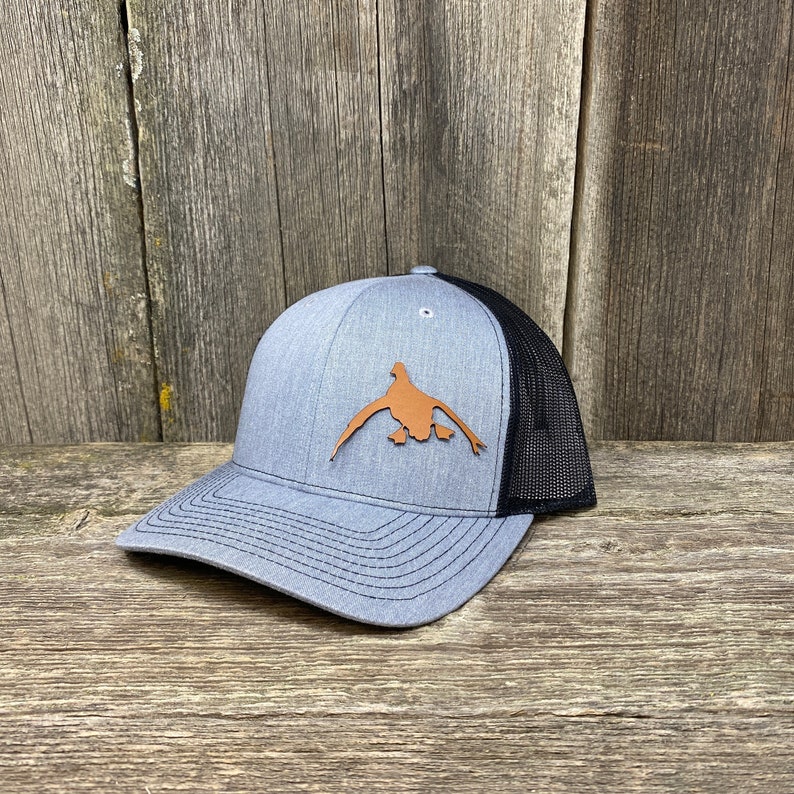 May include: A gray and black trucker hat with a brown leather duck silhouette on the front.