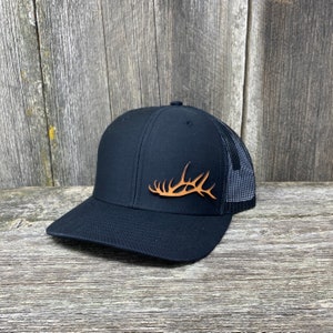 Może przedstawiać: Czarna czapka trucker z brązowym wzorem poroża łosia z przodu. Czapka ma czarny siateczkowy tył.