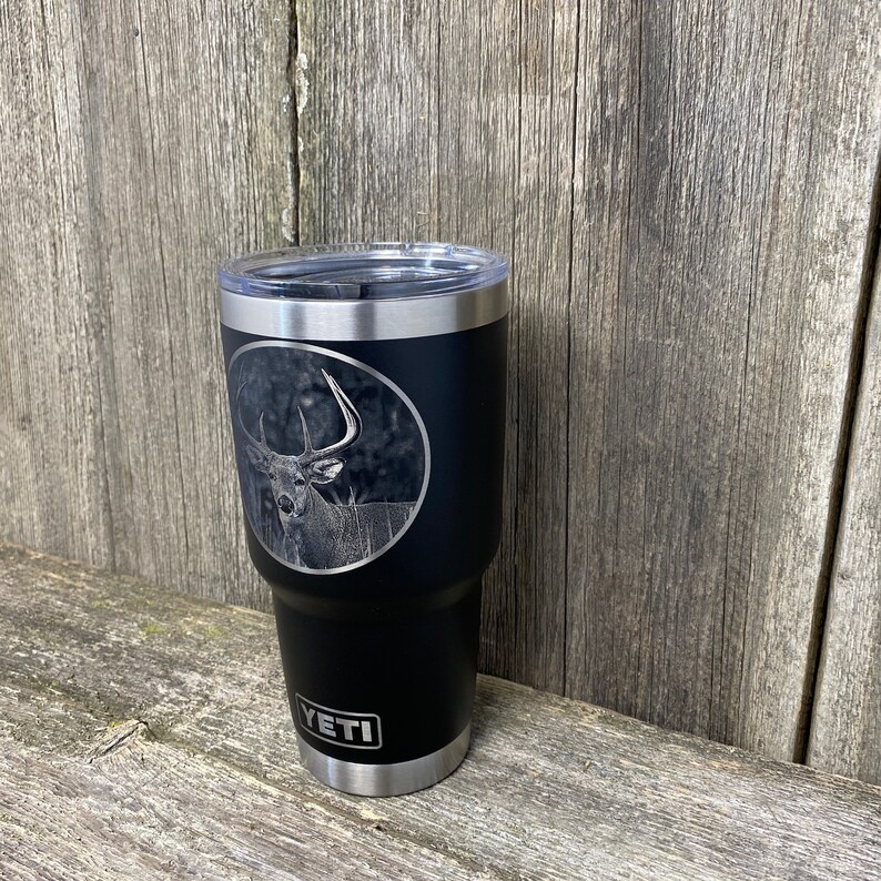 May include: A black Yeti tumbler with a stainless steel lid and base. The tumbler has a laser-etched design of a white-tailed deer with antlers.