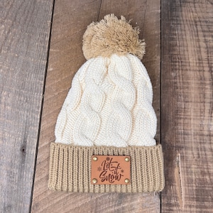 May include: White and brown knitted beanie with a pom pom on top. The beanie has a brown leather patch with the text "Let it Snow" on it.