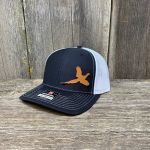 Może przedstawiać: Czarno-biała czapka trucker z brązową skórzaną naszywką w kształcie sylwetki bażanta z przodu.