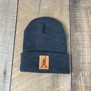 May include: A dark grey beanie with a brown leather patch featuring a silhouette of a Bigfoot. The patch has four brass rivets.