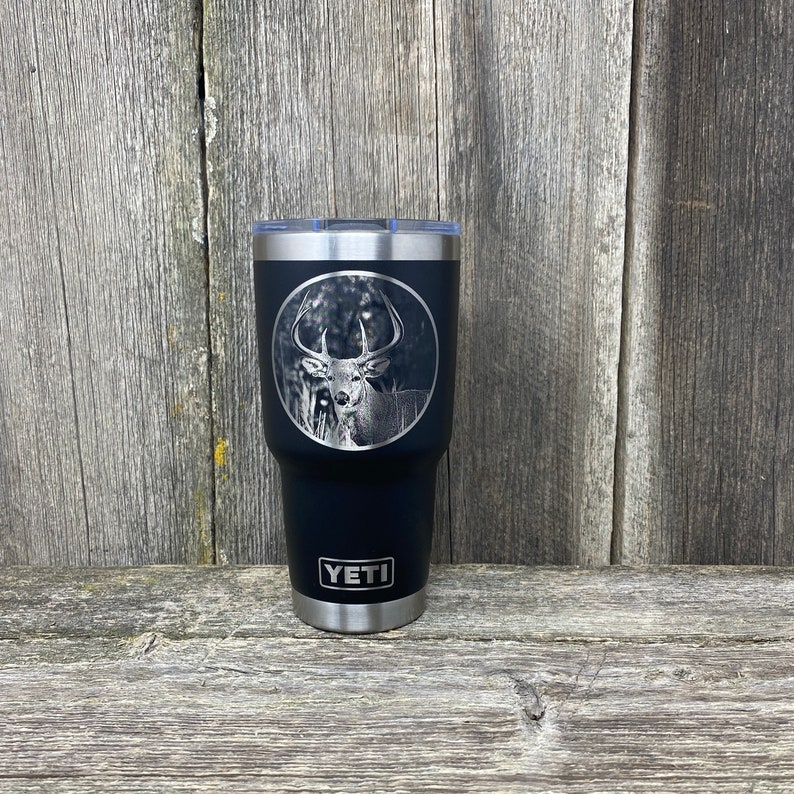 May include: Black Yeti tumbler with a laser engraved image of a white-tailed deer in a forest setting. The tumbler has the word "YETI" engraved on the bottom.