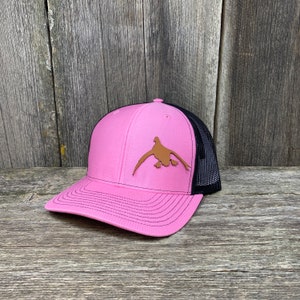 May include: Pink and black trucker hat with a brown leather duck silhouette on the front.