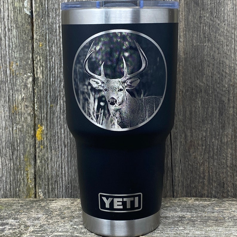 May include: Black Yeti tumbler with a laser engraved image of a white-tailed deer in a forest setting. The tumbler has the word "YETI" printed on the bottom.