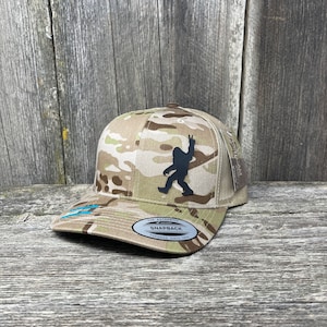 Könnte beinhalten: Eine Camouflage-Trucker-Kappe mit einer schwarzen Silhouette eines Bigfoot-Wesens, das den Friedenszeichen gibt. Die Kappe hat ein Netzrücken und ein braunes Leder-Patch mit der Aufschrift "The Authentic Snapback Trucker Since 1974."