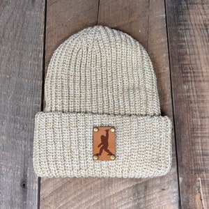 Pode incluir: Um gorro de malha bege com um remendo de couro marrom com a silhueta de um Pé Grande. O remendo tem dois rebites de latão.