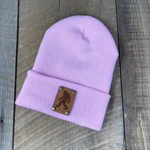 Puede incluir: Un gorro de color lila claro con un dobladillo. Un parche de cuero marrón con una silueta de Bigfoot está sujeto al dobladillo con remaches de latón. El gorro está hecho de un material de punto suave.