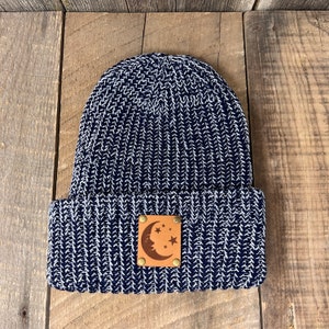 May include: A navy blue and white knitted beanie with a brown leather patch featuring a crescent moon and stars.