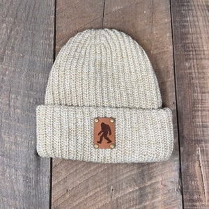 Op de afbeelding: Een beige gebreide beanie met een bruine leren patch met een silhouet van Bigfoot. De patch heeft messing klinknagels.