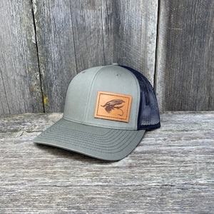May include: A green and black trucker hat with a brown leather patch featuring a fly fishing lure design.