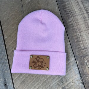 Puede incluir: Un gorro de punto rosa con un parche de cuero marrón que dice "Let it Snow" en letras blancas.