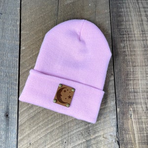 Könnte beinhalten: Eine hellviolette Beanie-Mütze mit umgeschlagenem Saum. Ein braunes Lederpatch mit einem Mond- und Stern-Design ist mit vier Messingnieten am Saum befestigt. Die Mütze ist aus einem weichen, gestrickten Material gefertigt.