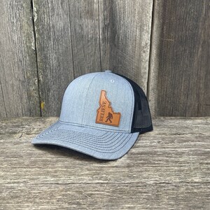 Könnte beinhalten: Eine graue und schwarze Trucker-Kappe mit einem braunen Leder-Patch, der den Umriss des Bundesstaates Idaho und eine Bigfoot-Silhouette zeigt. Der Patch enthält auch das Wort "BELIEVE".