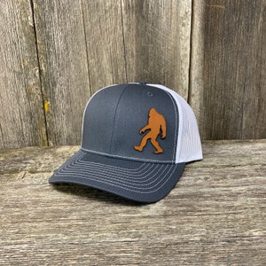 Könnte beinhalten: Eine dunkelgraue und weiße Trucker-Kappe mit einem braunen Leder-Bigfoot-Silhouetten-Patch auf der Vorderseite.