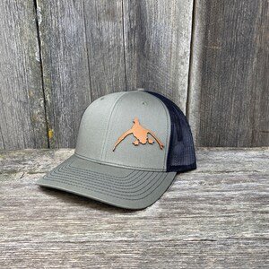 May include: A green and black trucker hat with a brown leather duck silhouette patch on the front.