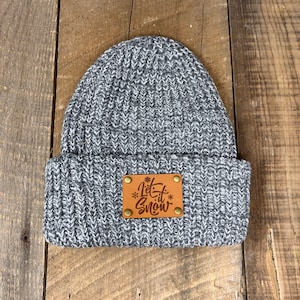 Puede incluir: Gorro de punto gris con un parche de cuero marrón que dice "Let it Snow" en letra blanca.