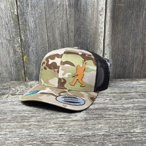 Könnte beinhalten: Eine Camouflage-Trucker-Kappe mit einem braunen Leder-Bigfoot-Patch. Die Kappe hat ein schwarzes Netzrücken und ein braunes Schild. Die Kappe hat den Text "The Authentic Snapback" und "Trucker Since 1978" auf der Rückseite.
