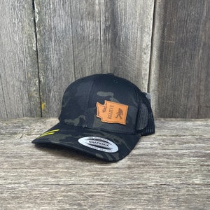 Könnte beinhalten: Schwarze und grüne Camouflage-Trucker-Kappe mit einem braunen Leder-Patch, der den Umriss des Bundesstaates Washington und die Worte "Believe" sowie eine Bigfoot-Silhouette zeigt.