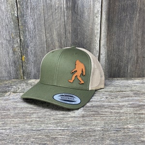 Könnte beinhalten: Olivgrüne und beige Trucker-Cap mit einem Lederaufnäher mit einer Bigfoot-Silhouette. Die Kappe hat einen gebogenen Schirm und eine Mesh-Rückseite. Der Aufnäher ist hellbraun. Auf der Vorderseite steht der Text "THE AUTHENTIC SNAPBACK TOPFONG SINCE 1974".