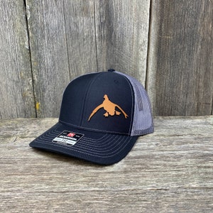 May include: A black and grey trucker hat with a brown leather duck silhouette on the front. The hat has a mesh back and a black adjustable strap.