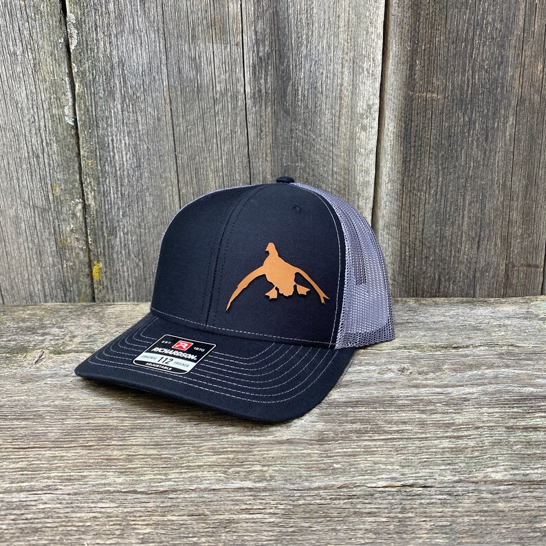 May include: A black and grey trucker hat with a brown leather duck silhouette on the front. The hat has a mesh back and a black adjustable strap.