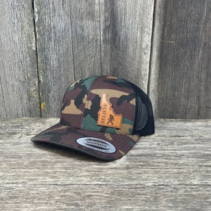 Könnte beinhalten: Camouflage-Baseballkappe mit schwarzem Mesh-Rücken. Ein Lederaufnäher zeigt die Umrisse von Idaho, das Wort "BELIEVE" und eine Bigfoot-Silhouette. Die Kappe hat einen gebogenen Schirm und einen Snapback-Verschluss.
