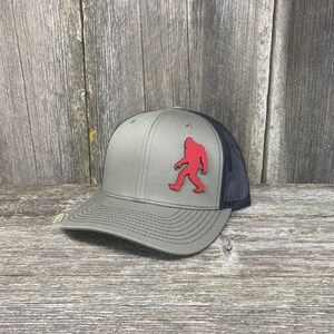 Könnte beinhalten: Eine grüne und schwarze Trucker-Kappe mit einer roten Bigfoot-Silhouette auf der Vorderseite.