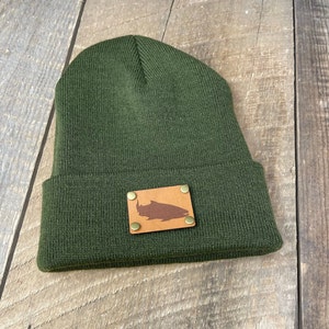 May include: A dark green knitted beanie with a brown leather patch featuring a silhouette of a fish. The patch has four gold rivets.