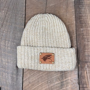 May include: A beige knitted beanie with a brown leather patch featuring a fly silhouette.