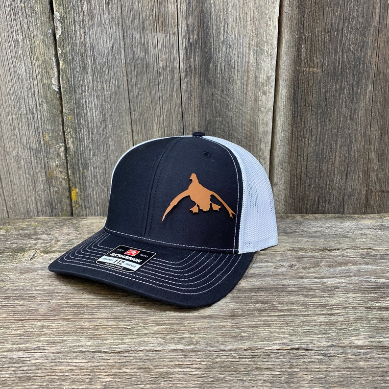 May include: A black and white trucker hat with a brown leather duck silhouette patch. The hat has a mesh back and a black brim.
