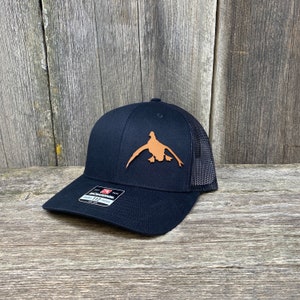 May include: A black and brown trucker hat with a brown leather duck silhouette on the front. The hat has a black mesh back.