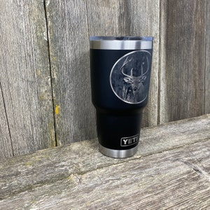 May include: Black Yeti tumbler with a laser engraved image of a whitetail deer in a circle. The tumbler has the word "YETI" printed on the bottom.