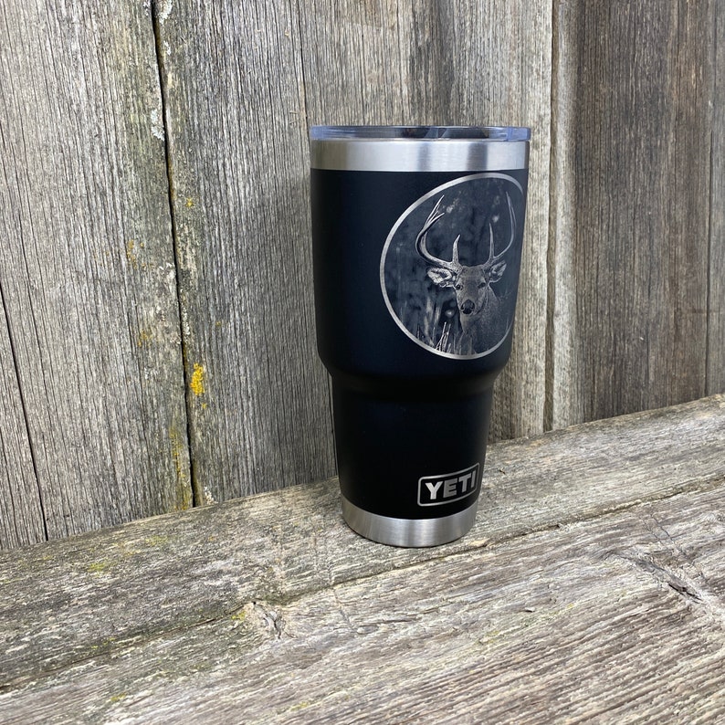 May include: Black Yeti tumbler with a laser engraved image of a whitetail deer in a circle. The tumbler has the word "YETI" printed on the bottom.