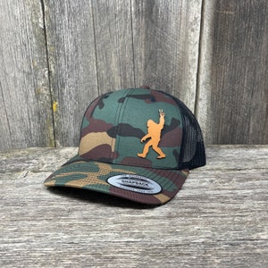 Könnte beinhalten: Eine Camouflage-Trucker-Kappe mit einem braunen Leder-Bigfoot-Patch. Der Patch zeigt Bigfoot mit einem Peace-Zeichen. Die Kappe hat ein schwarzes Mesh-Rückteil und einen schwarzen Druckknopfverschluss. Die Kappe hat einen Aufkleber auf der Vorderseite, auf dem "The Authentic Snapback Supplying Since 1974" steht.