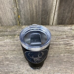 May include: Black Yeti tumbler with a laser engraved design of a white-tailed deer in a forest setting. The tumbler has a black lid with a silver band.