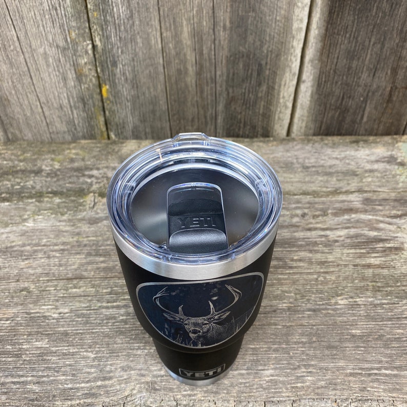 May include: Black Yeti tumbler with a laser engraved design of a white-tailed deer in a forest setting. The tumbler has a black lid with a silver band.