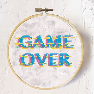 Op de afbeelding: Een witte borduurring met de woorden "GAME OVER" geborduurd in blauw, roze, geel en groen. De letters zijn gepixeld, wat een retro videogame-esthetiek geeft.