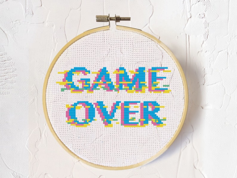 Op de afbeelding: Een witte borduurring met de woorden "GAME OVER" geborduurd in blauw, roze, geel en groen. De letters zijn gepixeld, wat een retro videogame-esthetiek geeft.