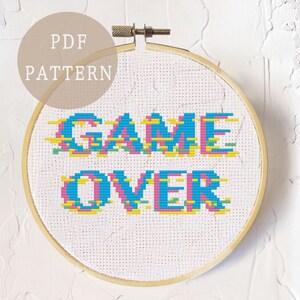 Op de afbeelding: Een kruissteekpatroon met de tekst "GAME OVER" in een gepixelde stijl met blauwe, roze, gele en groene kleuren. Het patroon wordt weergegeven in een houten borduurring.