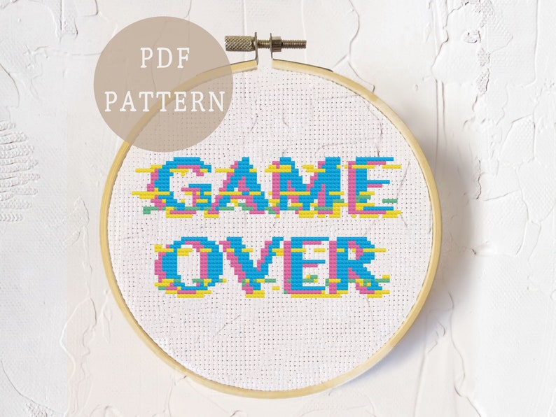 Op de afbeelding: Een kruissteekpatroon met de tekst "GAME OVER" in een gepixelde stijl met blauwe, roze, gele en groene kleuren. Het patroon wordt weergegeven in een houten borduurring.