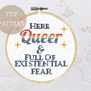 May include: A white embroidery hoop with a rainbow-coloured word "Queer" and the text "Here Queer & Full of Existential Fear".