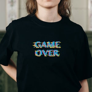 Op de afbeelding: Zwart T-shirt met een gepixelde "GAME OVER" afbeelding in blauw, roze en geel.